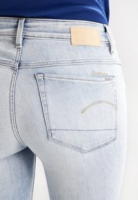 Vaqueros de mezclilla azul claro con una cintura de altura media, bolsillos traseros con costuras sutiles y una etiqueta de marca de cuero en la cinturilla.