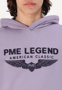 Svetlofialová mikina s čiernym vyšitým logom s krídlami. Dizajn obsahuje prominentný nápis "PME LEGEND AMERICAN CLASSIC" na prednej strane.