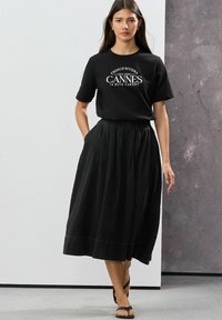 Tricou din bumbac negru cu imprimeu alb "French Riviera Cannes", asortat cu o fustă lungă plisată neagră, având un tiv lat și buzunare.