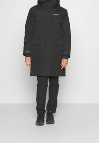 Parka aislante negra con capucha, que presenta dos bolsillos frontales y puños elásticos. Llevada con pantalones oscuros a juego y zapatillas negras.