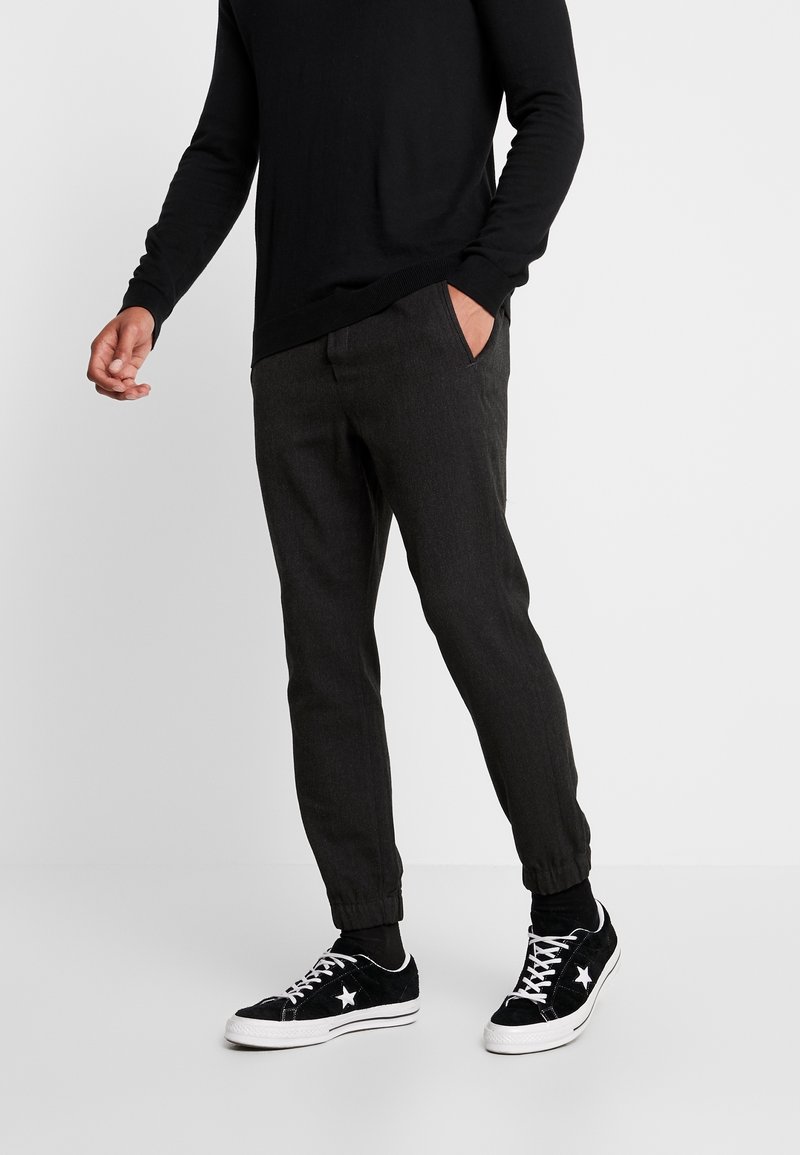 Tummanharmaat kapenevat housut, joissa on sivutaskut, yhdistettynä mustiin lenkkareihin, joissa on valkoisia tähtiä. Kevyt kangas, jossa on sileä rakenne.