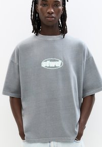 Graues, übergroßes T-Shirt mit rundem Halsausschnitt und kurzen Ärmeln, mit einem mintgrünen Logo "stwd", das auf der Brust zentriert ist. Glatte Baumwolltextur.