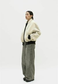 Cremfarbene Kunstfelljacke mit schwarzem Rippbündchen, kombiniert mit grauen, hoch tailliertenweiten Jeans und schwarzen Plateauschuhen, vor einem weißen Hintergrund.