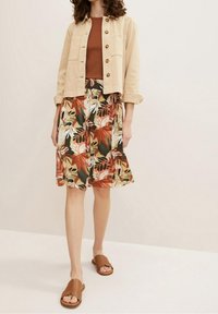 Veste beige courte avec des boutons marron, débardeur rouille, et jupe fleurie aux motifs orange, vert et blanc, associée à des sandales à enfiler marron.