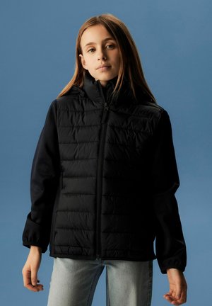 Ung person med langt hår iført en sort quiltet jakke og lyseblå jeans mod en ensfarvet blå baggrund.