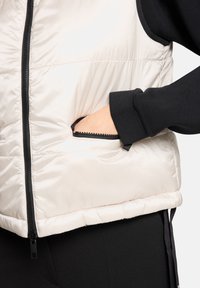 Jasnobeżowa pikowana kamizelka z zamkiem błyskawicznym z przodu, wyposażona w boczne kieszenie i czarne podszycie. Gładka, błyszcząca faktura z elastycznymi akcentami.