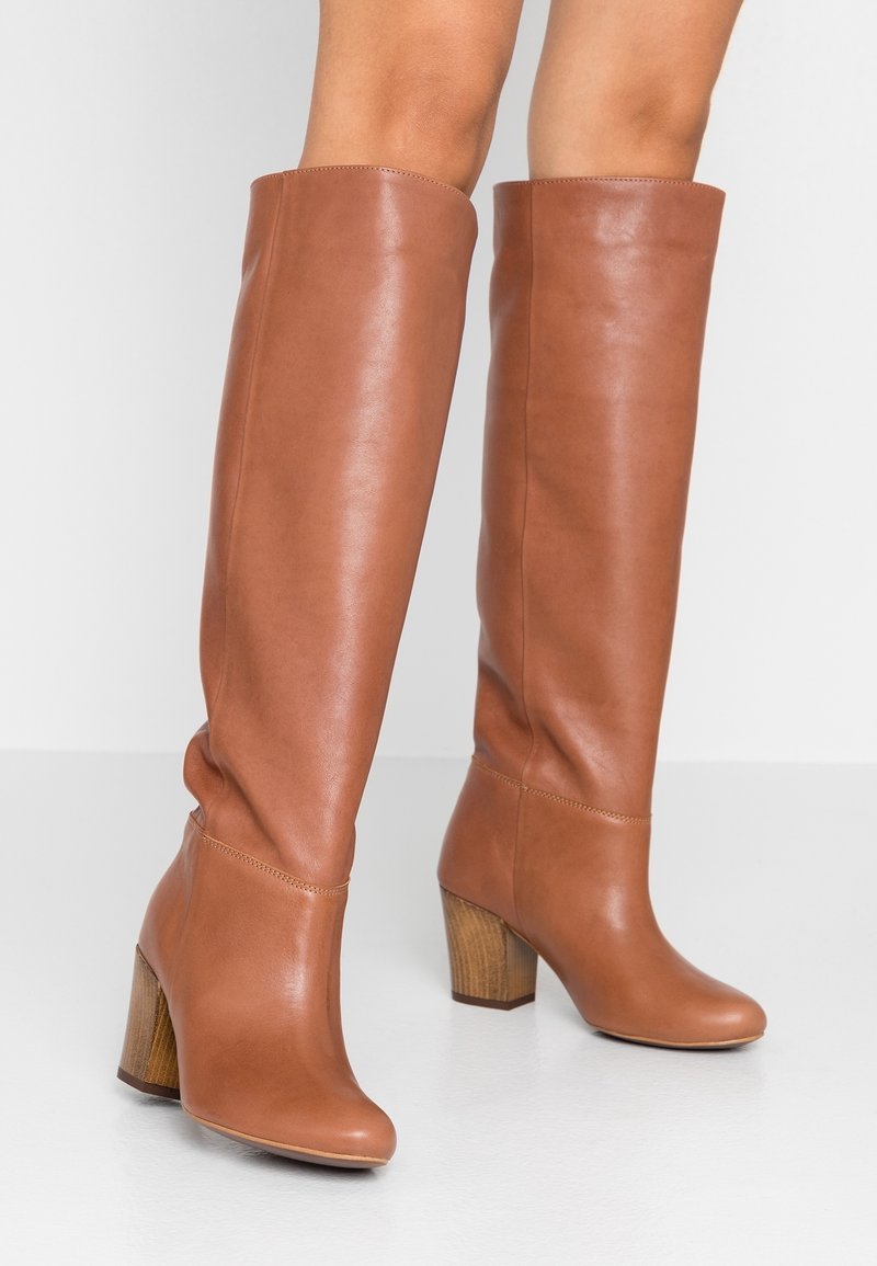 Bottes hautes en cuir marron clair avec talons en bois blocs portées sur des jambes nues sur un fond blanc.