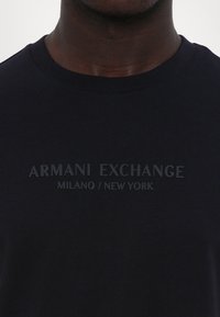 Tamsiai mėlynas apvalaus kaklo marškinėliai, pagaminti iš medvilnės, su matinės spaudos logotipu "ARMANI EXCHANGE MILANO / NEW YORK" priekyje.