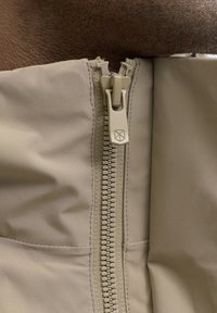 Beiger Reißverschluss mit ineinandergreifenden Zähnen, an Stoff ähnlicher Farbe befestigt. Verfügt über einen markengeprägten Reißverschlussanhänger und eine glatte Textur, teilweise geöffnet.