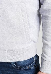 Felpa grigia realizzata in un tessuto morbido e testurizzato, con tasca laterale e orlo a costine. Indossata sopra jeans blu scuro con cuciture visibili.