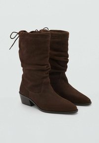 Botas de tobillo de ante marrón con un diseño holgado, puntas afiladas y un tacón apilado. Presentan una parte superior fruncida con un cierre de lazo para un ajuste ajustable.