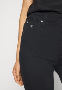 Jeans de mezclilla negra con un diseño ajustado, que cuentan con herrajes plateados, bolsillos frontales y un logo bordado sutil en la cinturilla.