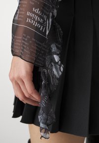 Průhledná černá blouse s potištěným textem, dlouhými rukávy a volánkovými okraji, v kombinaci s černou plisovanou sukni. Ruka je viditelná, jak drží blouse.