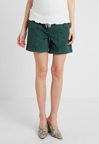 Green cotton shorts with a relaxed fit, feature a drawstring waist, and contrasting stitching. Paired with patterned heeled shoes.