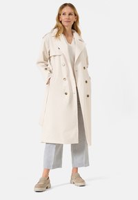 Manteau trench beige clair à double boutonnage avec de gros boutons, large col, poches latérales et design mi-long, associé à un pantalon gris et des chaussures.