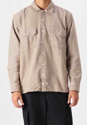 Mand iført beige og hvid ternet skjorte med knapper og to brystlommer samt sorte bukser mod en ensfarvet baggrund.