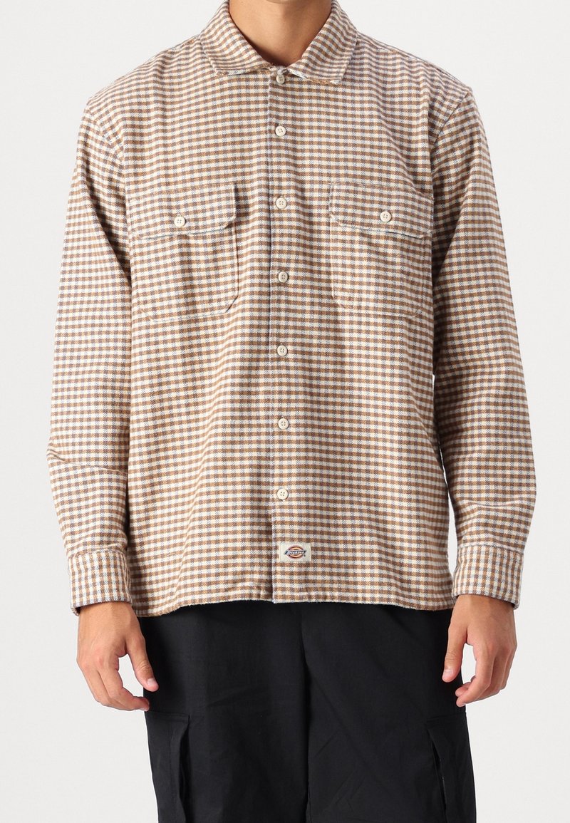 Homme portant une chemise à carreaux beige et blanche boutonnée avec deux poches poitrine et un pantalon noir sur un fond uni.