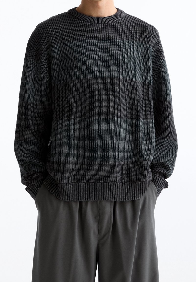Homme portant un pull en maille à côtes rayé noir et gris foncé, les mains dans les poches d'un pantalon ample gris foncé sur fond blanc.