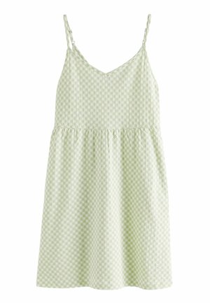 Femme portant une robe d'été vichy vert clair et blanc à fines bretelles, debout à l'extérieur près d'un bâtiment en plein soleil.