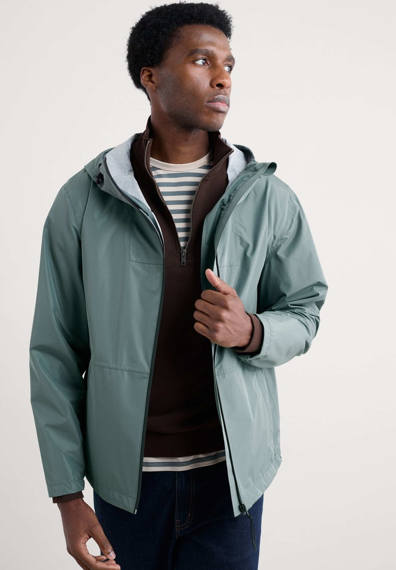 Giacca impermeabile verde chiaro con cappuccio, foderata di grigio, con tasche con zip, indossata sopra un maglione marrone con zip e una maglietta a righe, abbinata a jeans scuri.