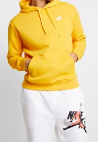 Sudadera amarilla con un bolsillo frontal, capucha con cordón y un pequeño logo blanco de Nike en el pecho. Combinada con pantalones blancos con detalles gráficos.