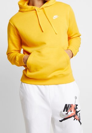 Geel hoodie met een voorzak, een capuchon met trekkoord en een klein wit Nike-logo op de borst. Gepaard met witte broek met grafische details.