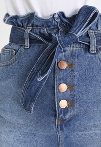 Džínová sukně s vysokým pasem, která má volánkový pás, látkový pásek a tři bronzové knoflíky na zapínání. Modrý denim s mírným vyblednutím.