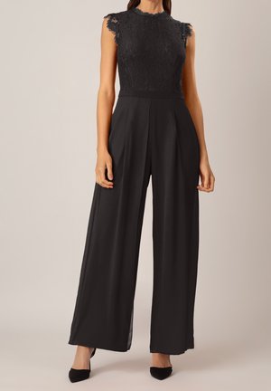 Femme portant une combinaison noire en dentelle sans manches avec un pantalon large et des talons noirs sur un fond uni.