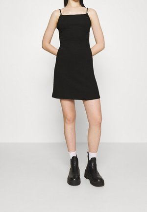 Vrouw in een zwarte mouwloze minijurk met witte sokken en zwarte enkellaarsjes, staand tegen een effen witte achtergrond.