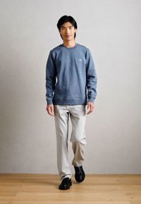 Sweatshirt cinza com um pequeno bolso no peito, calças de cor clara e sapatos pretos. O modelo está em pé sobre um piso de madeira contra um fundo neutro.