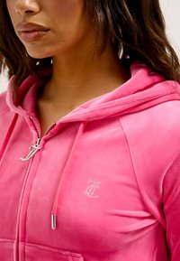 Roze velours zip-up hoodie met een zilveren ritssluiting en geborduurd logo met rhinestones, met een gladde textuur en capuchon ontwerp.