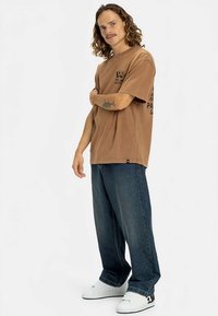 Hnědé tričko s krátkými rukávy s grafickým potiskem, doplněné volnými tmavými džínami a bílými teniskami. Tričko má pohodlný střih a zaoblený spodní lem.