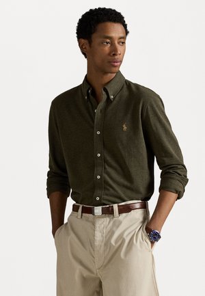 Camicia verde oliva con bottoni, maniche lunghe e un piccolo logo. Abbinata a pantaloni beige e una cintura marrone. Orologio con quadrante blu.