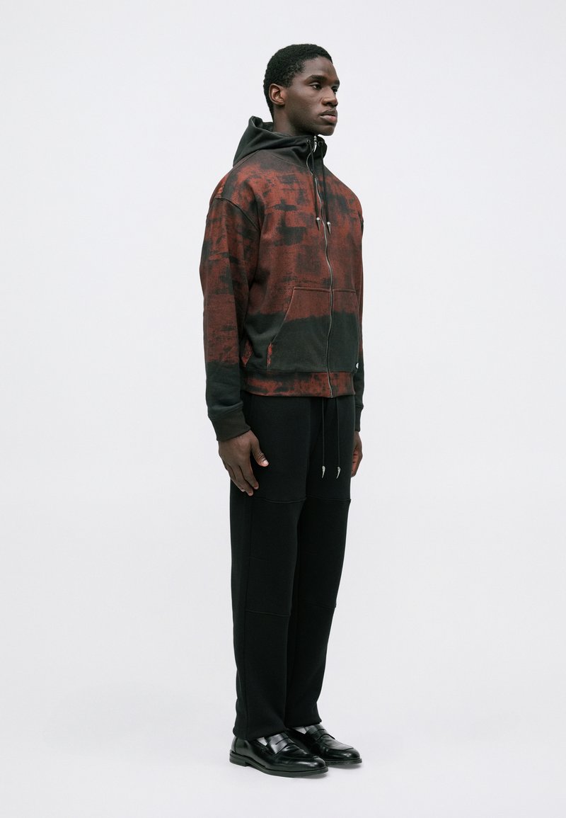 Ung mand står iført en rød og sort hættetrøje med sorte bukser og sorte læder sko, vendt sidelæns mod en hvid baggrund.