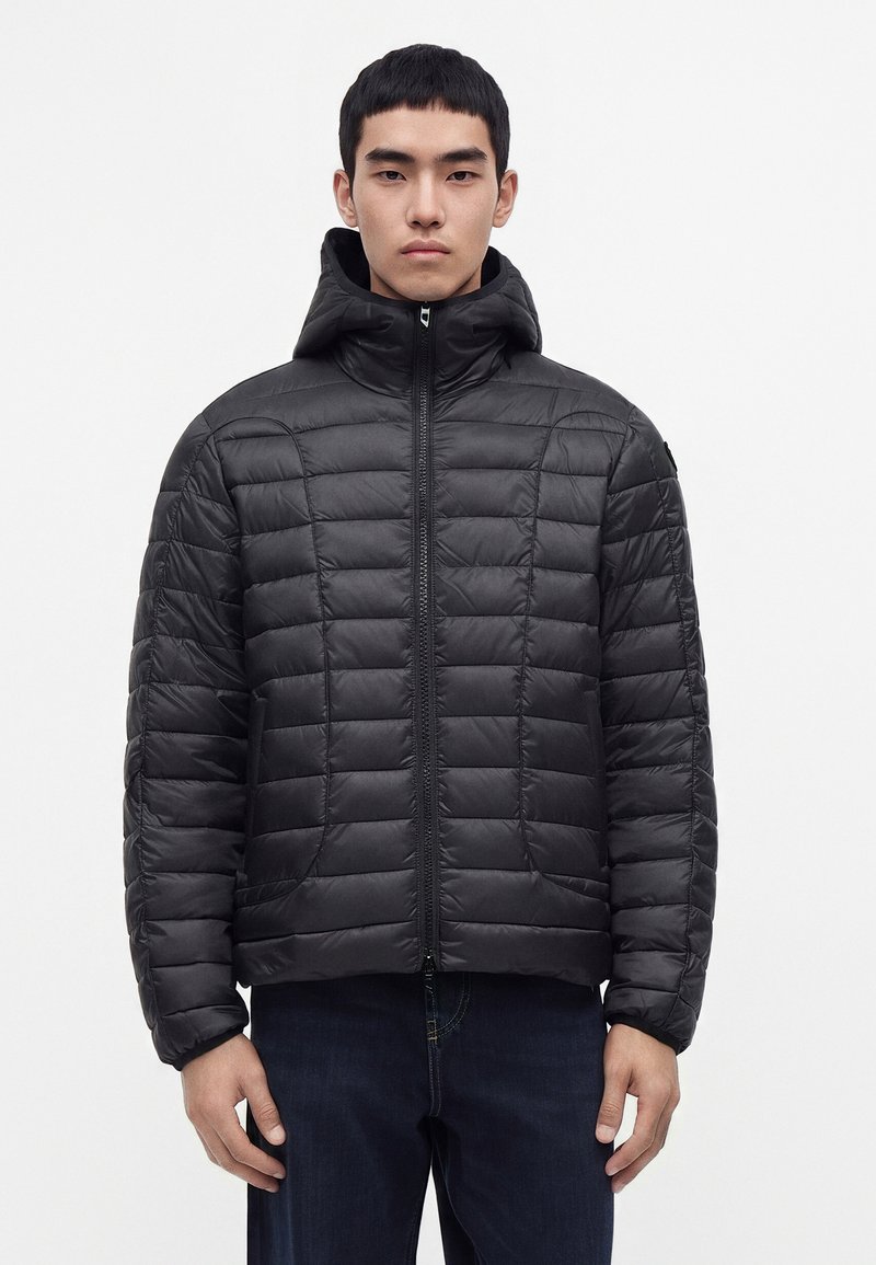 Jeune homme portant une veste matelassée noire à capuche, zipée, debout droit devant un fond blanc uni.