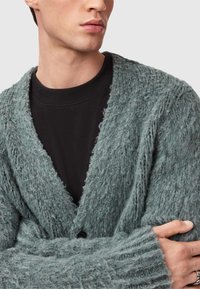 Teksturowany, oversize kardigan w jasnym odcieniu teal, z głębokim dekoltem w serek, zapięciem na guziki oraz grubym wzorem dzianiny, noszony z czarną bluzą z okrągłym dekoltem pod spodem.