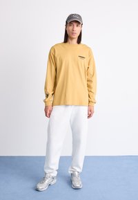 Chemise à manches longues jaune avec logo noir sur la poitrine, associée à un pantalon de survêtement blanc et des chaussures de sport argentées, complétée par une casquette grise.