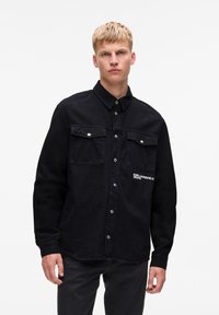 Plavi muškarac u crnoj košulji na dugme s džepovima na prsima i logom "Karl Lagerfeld Jeans" s lijeve strane, stoji protiv bijele pozadine.