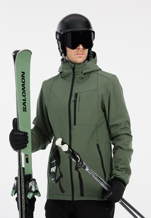 Grøn vandtæt skijakke med lynlås på forsiden, hætte og brystlomme, parret med sorte skihandsker, hjelm og ski med sorte og grønne accenter.