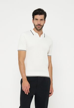 Homme portant un polo en maille blanche à manches courtes avec bordure noire au col, aux manches et à l'ourlet, associé à un pantalon noir, debout sur un fond blanc.