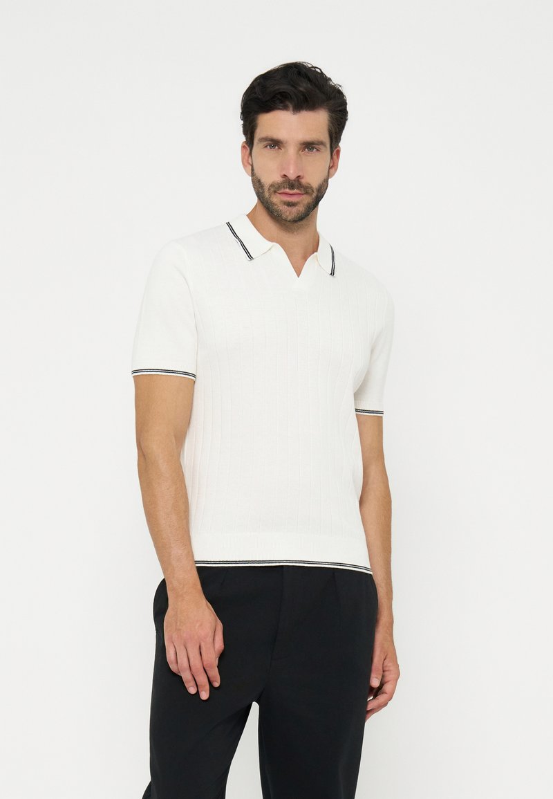Homme portant un polo en maille blanche à manches courtes avec bordure noire au col, aux manches et à l'ourlet, associé à un pantalon noir, debout sur un fond blanc.