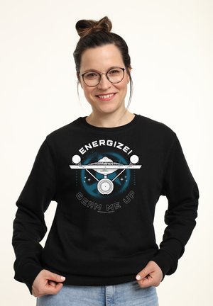Smilende kvinde med briller iført en sort sweatshirt med et rumskib og teksten "Energize! Beam me up."