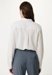 Blusa bianca a maniche lunghe con colletto classico, dettaglio sulla schiena, e texture liscia, abbinata a pantaloni houndstooth blu e bianchi.