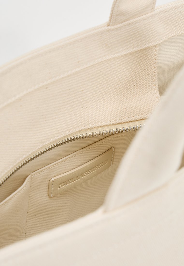 Sac fourre-tout en toile beige avec un compartiment zippé, présentant une étiquette en cuir lisse estampillée "KARL LAGERFELD" à l'intérieur.