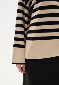 Pulover striped dintr-un amestec de lână bej și neagră, cu benzi late, mâneci lungi și o croială lejeră, expus împreună cu o parte dintr-o fustă neagră.