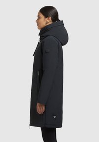 Tmavomodrá parka s kapucňou, ktorá má zaoblený golier, zips, bočné vrecká a nášivku na leme. Hladká textúra látky.