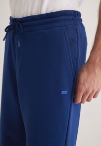 Navyblaue Sweatpants mit einem Kordelzugbund, ausgestattet mit seitlichen Taschen und einem kleinen blauen Logosticker am Oberschenkel. Weicher Stofftextur.