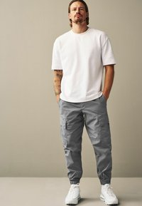 Tricou alb cu mânecă scurtă, pantaloni cargo gri cu buzunare și adidași albi. Ținuta are o croială lejeră și texturi moi.