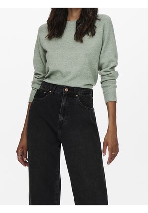 Femme portant un pull vert clair à manches longues rentré dans un jean noir taille haute à jambes larges, avec des bagues aux deux mains.