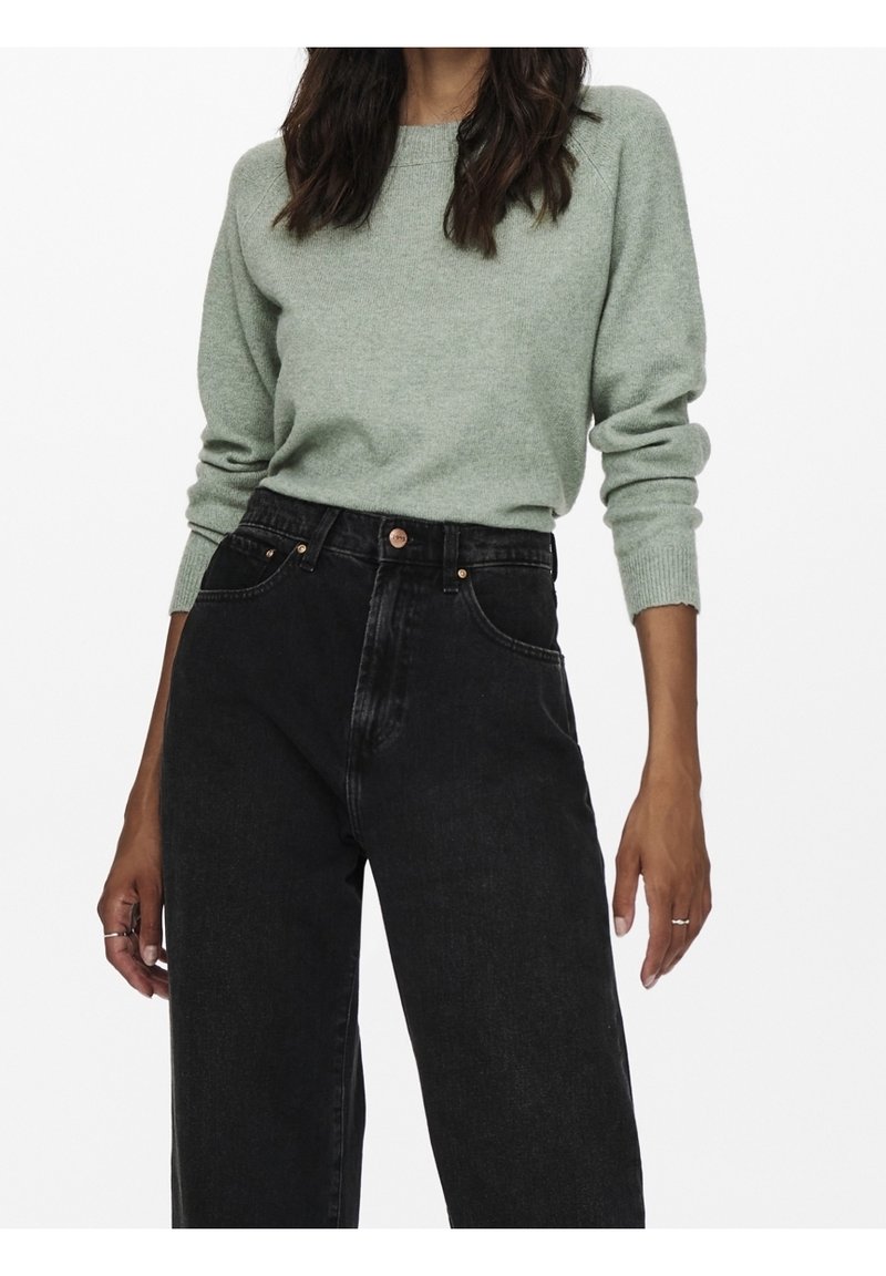 Femme portant un pull vert clair à manches longues rentré dans un jean noir taille haute à jambes larges, avec des bagues aux deux mains.